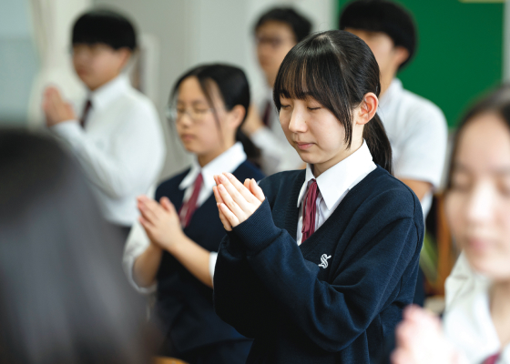 朝礼で合掌する生徒と校内の額